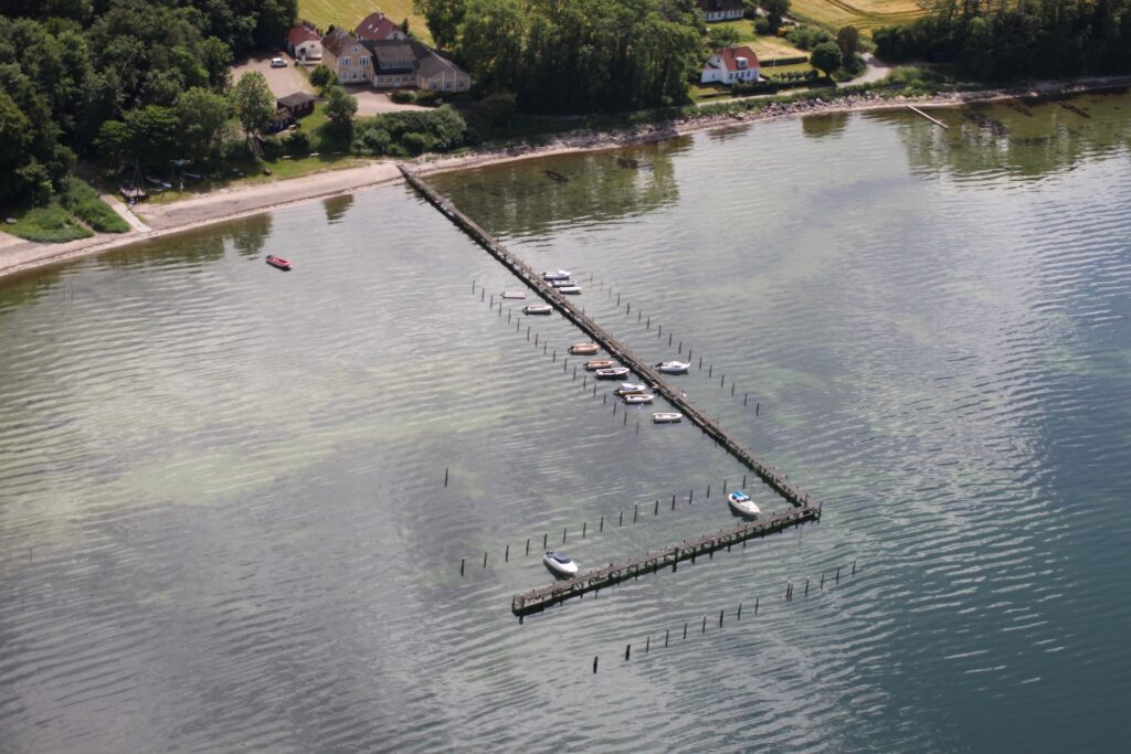 L-förmiger Holzsteg mit kleinen Booten an ruhiger Küstenbucht, dahinter Strand und vereinzelte Häuser.