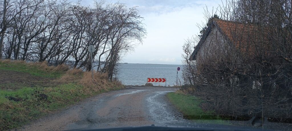 Matschiger Feldweg endet am See; rot-weiße Chevron-Sperre warnt, flankiert von kahlen Bäumen und Haus mit Ziegeldach