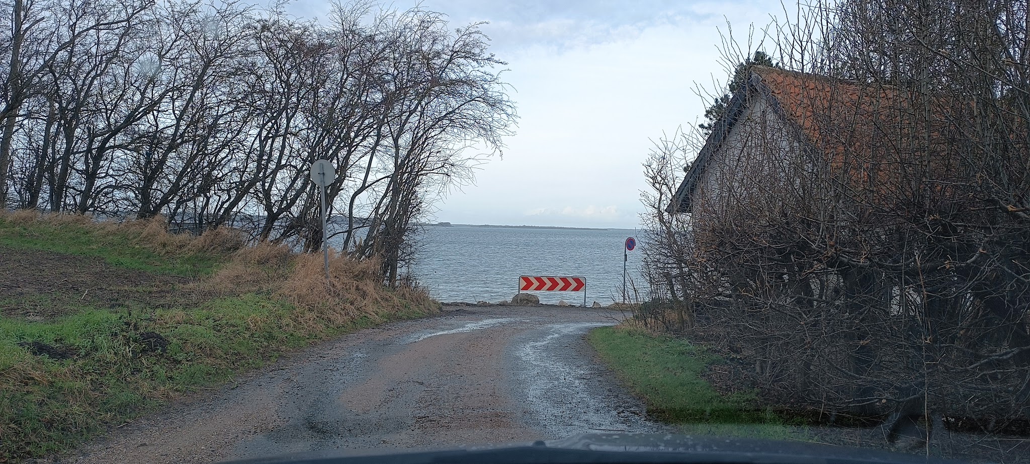 Matschiger Feldweg endet am See; rot-weiße Chevron-Sperre warnt, flankiert von kahlen Bäumen und Haus mit Ziegeldach
