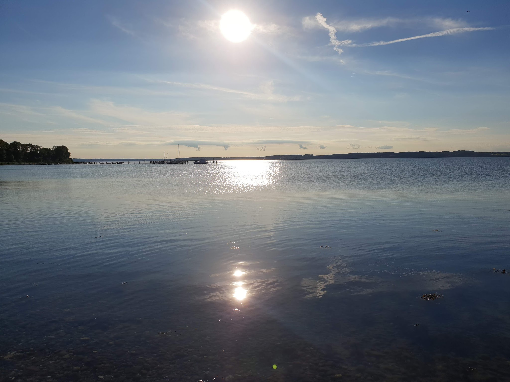 Tiefer Sonnenstand spiegelt sich auf ruhigem See, Steg mit Segelbooten vor ferner Uferlinie.