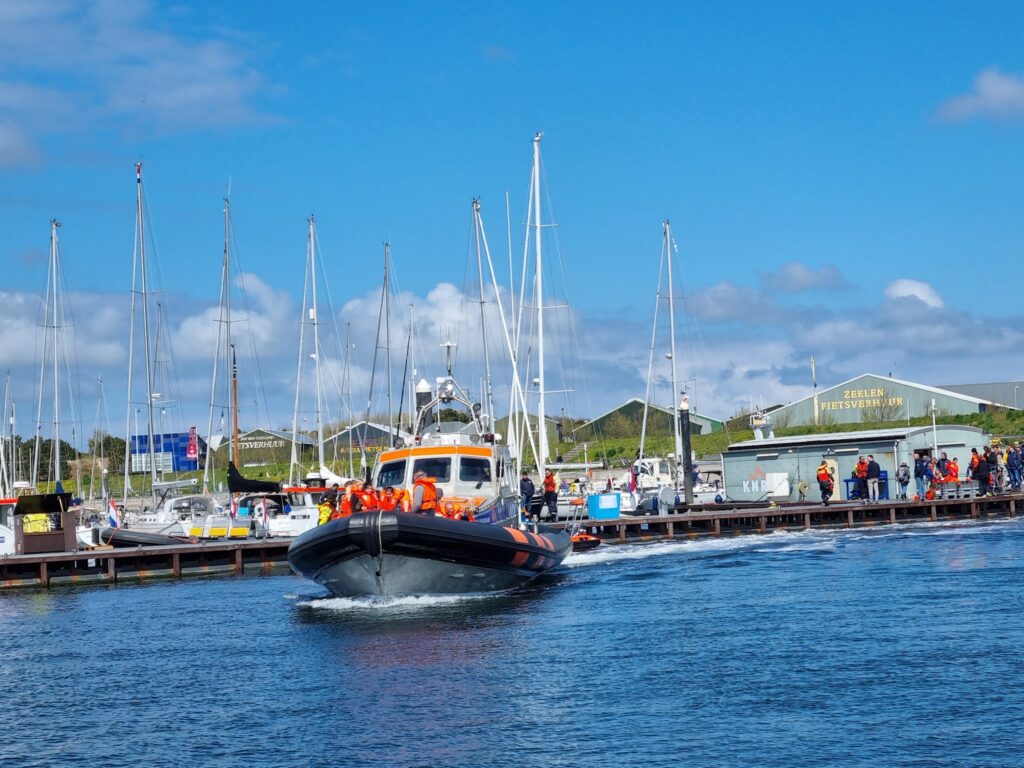 Rettungsboot mit orange gekleideten Passagieren fährt aus Jachthafen, Segelboote und Zuschauer auf dem Steg.