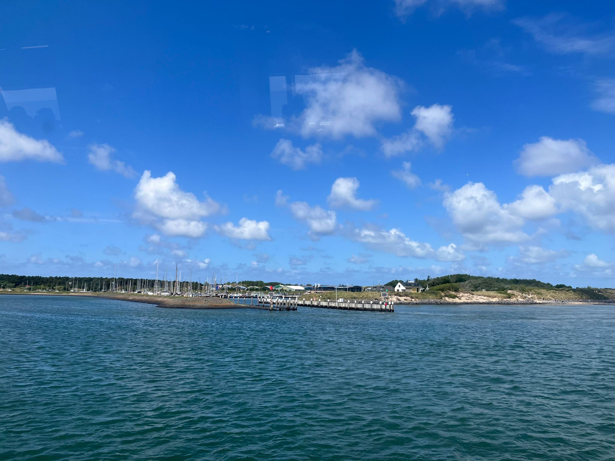 Küste mit kleinem Hafen: Reihen hoher Segelmasten, Holzpier, grünes Wasser unter blauem Himmel mit weißen Wolken.