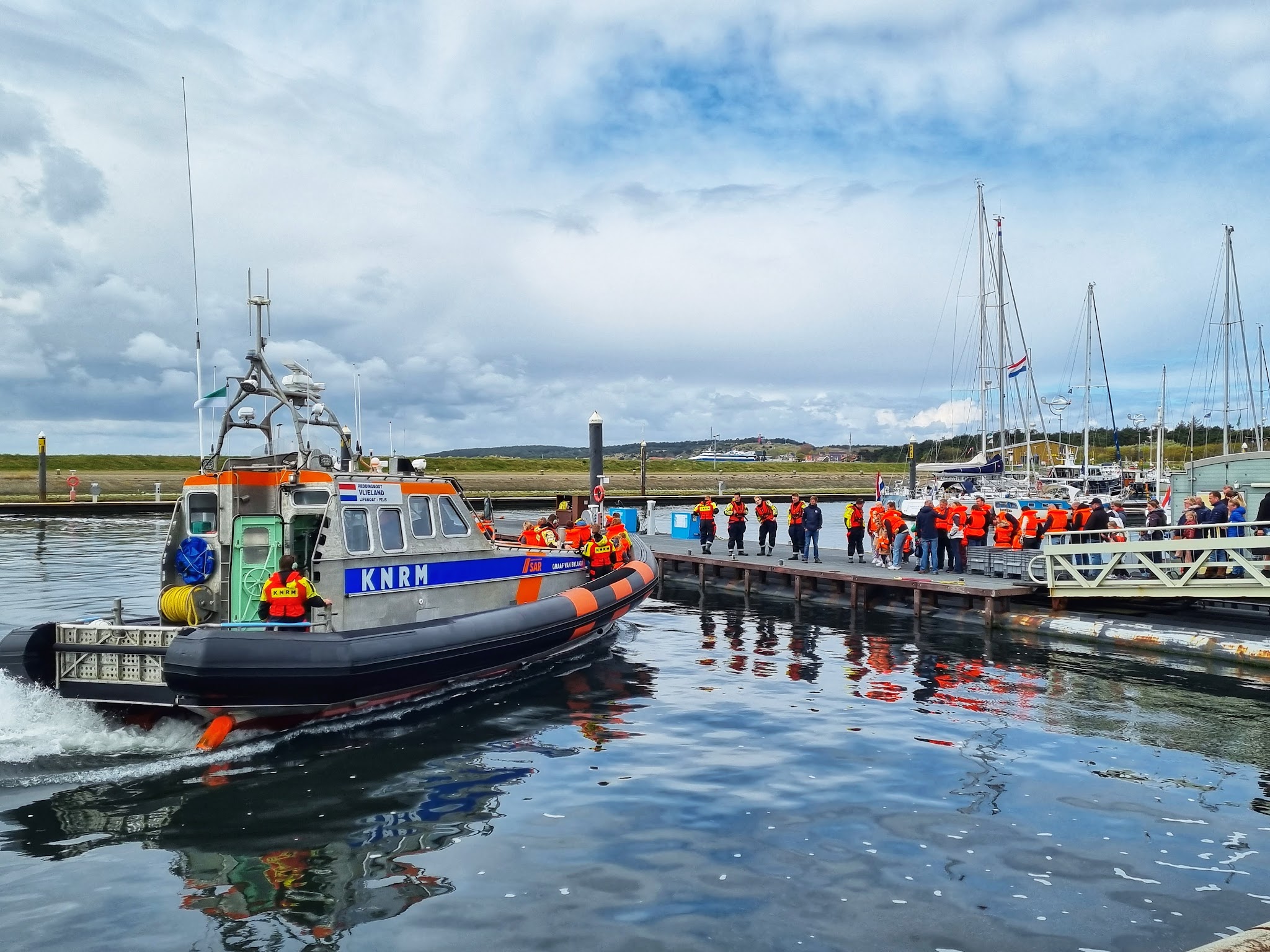 KNRM-Seenotrettungsboot legt ab, während zahlreiche Menschen in orange Rettungswesten am Steg im Hafen warten.