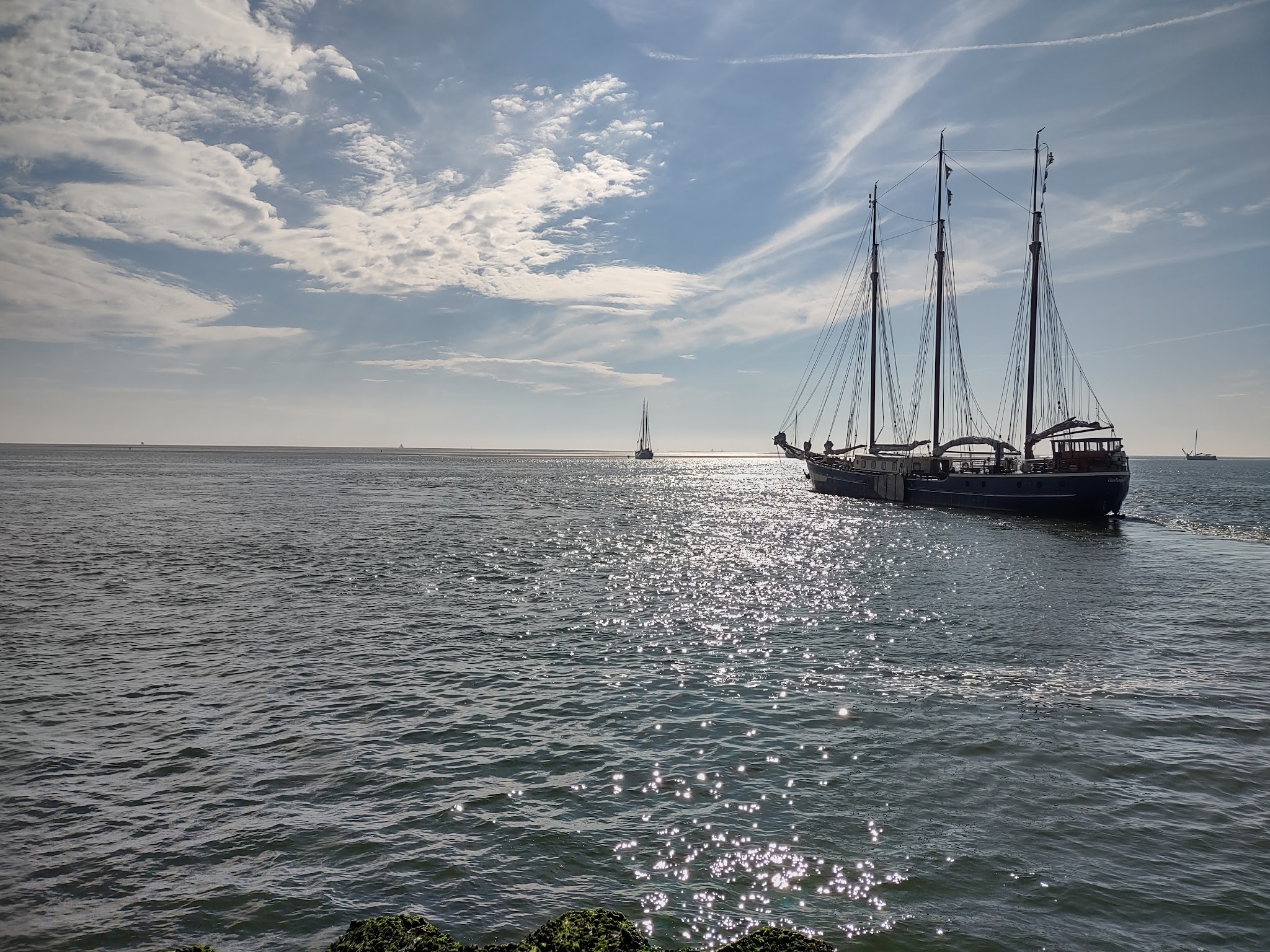 Dreimast-Segelschiff fährt über glitzerndes Meer; ferne Segelboote am Horizont unter teils bewölktem Himmel.