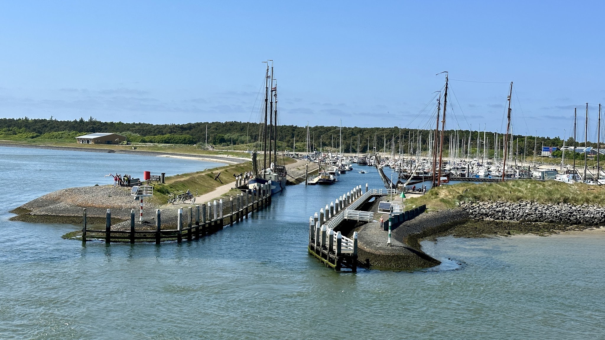 Schmale Hafeneinfahrt zwischen Grasdünen; zahlreiche Segelboote liegen an Stegen unter klarem blauen Himmel.