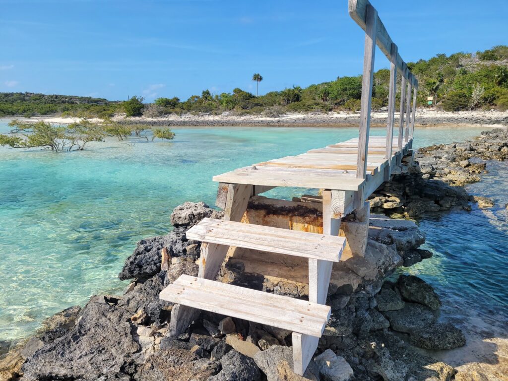 Schmale, alte Holzbrücke führt über türkisfarbenes, flaches Meer zu palmengesäumter Inselküste.