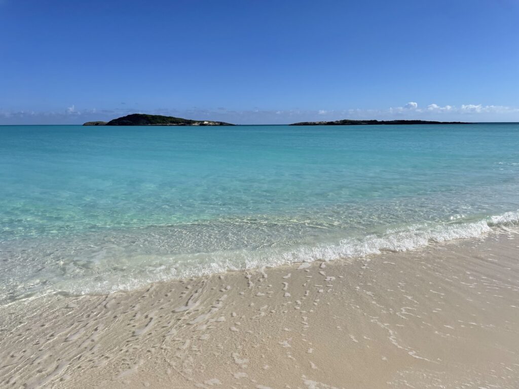 Türkises Meer mit sanften Wellen am hellen Sandstrand; am Horizont zwei grüne Inseln unter klarem blauen Himmel.