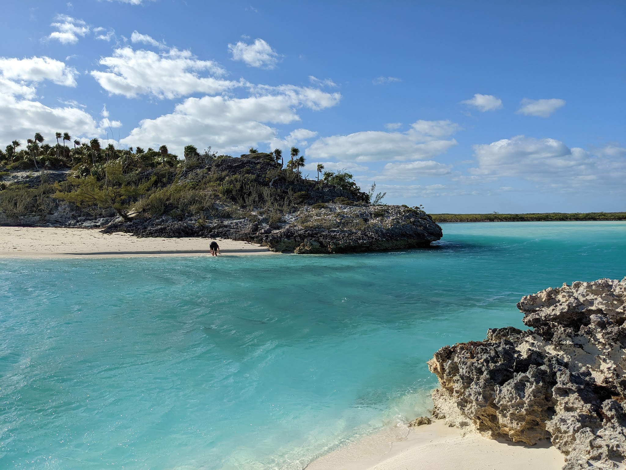 Türkisfarbene Lagune, weißer Strand mit Felsen, palmenbewachsene Klippe; einzelne Person kniet am Ufer.
