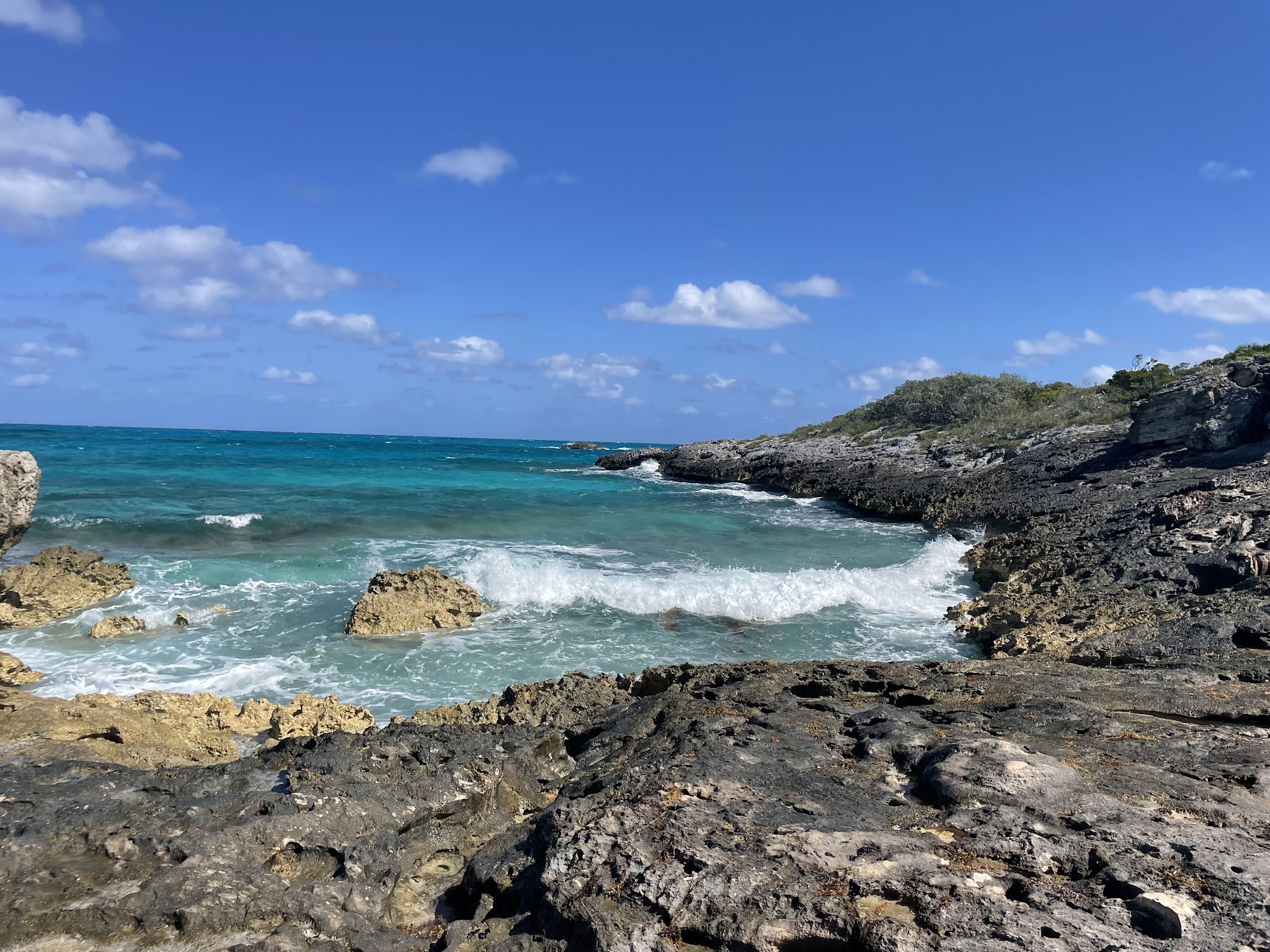 Türkisfarbene Wellen schlagen gegen dunkle, zerklüftete Felsküste unter wolkenlosem, tiefblauem Himmel.