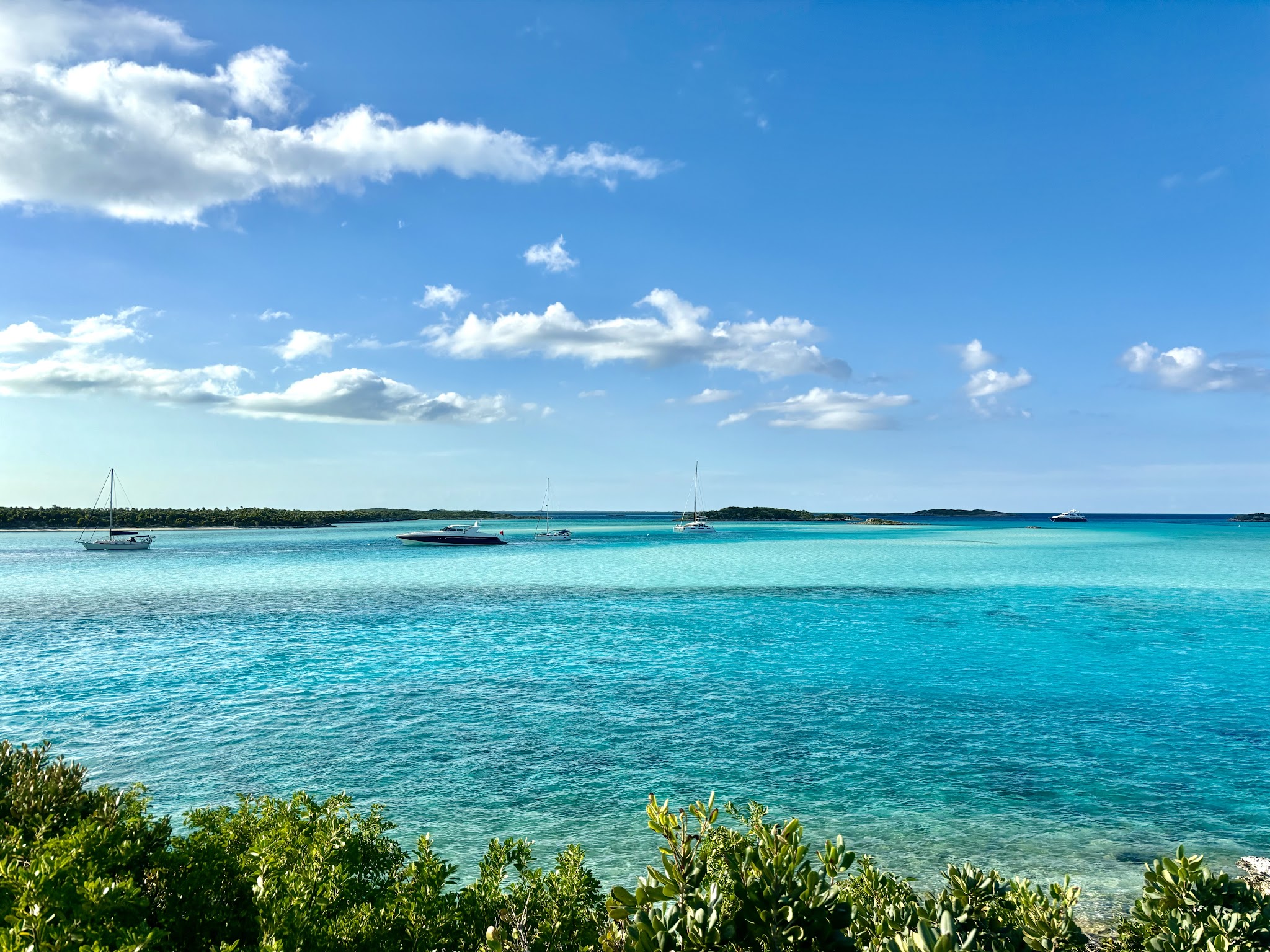 Türkises Meer mit drei Segelbooten vor flacher Insel, grüne Büsche im Vordergrund, blauer Himmel mit Wolken.
