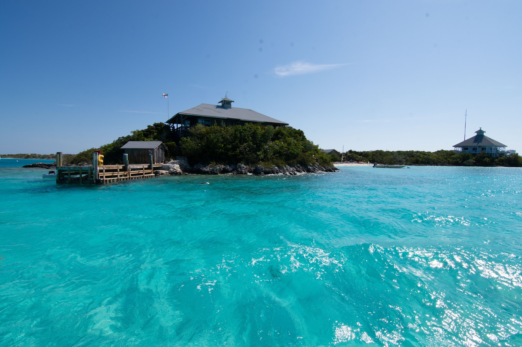 Türkises, klares Meer umspült kleine Insel mit Holzsteg, dichtem Grün und zwei Pfahlhäusern unter blauem Himmel.