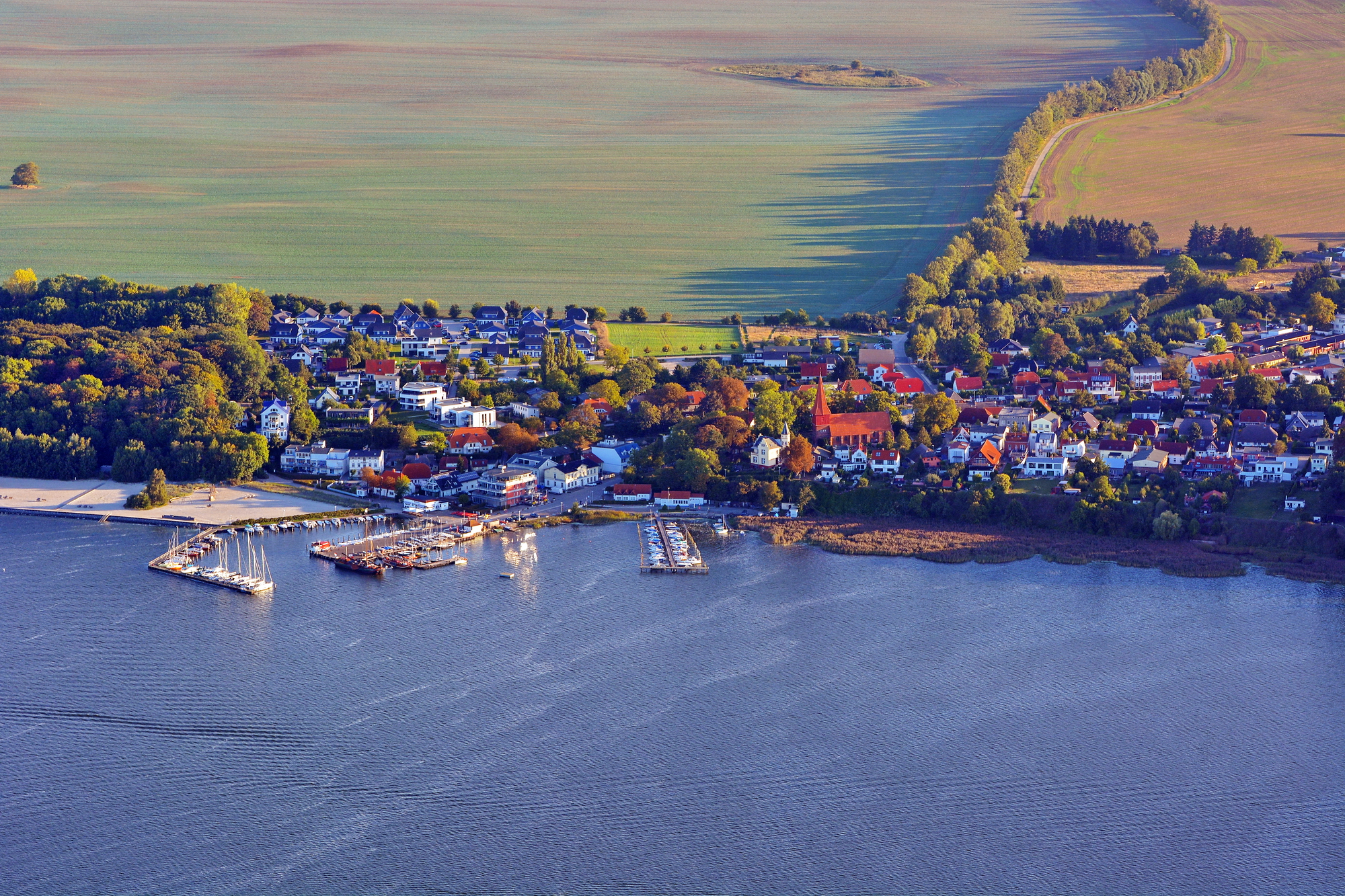 Kleines Küstendorf mit Yachthafen und Segelbooten, roter Kirchturm, von Feldern, Wald und Wasser umgeben.