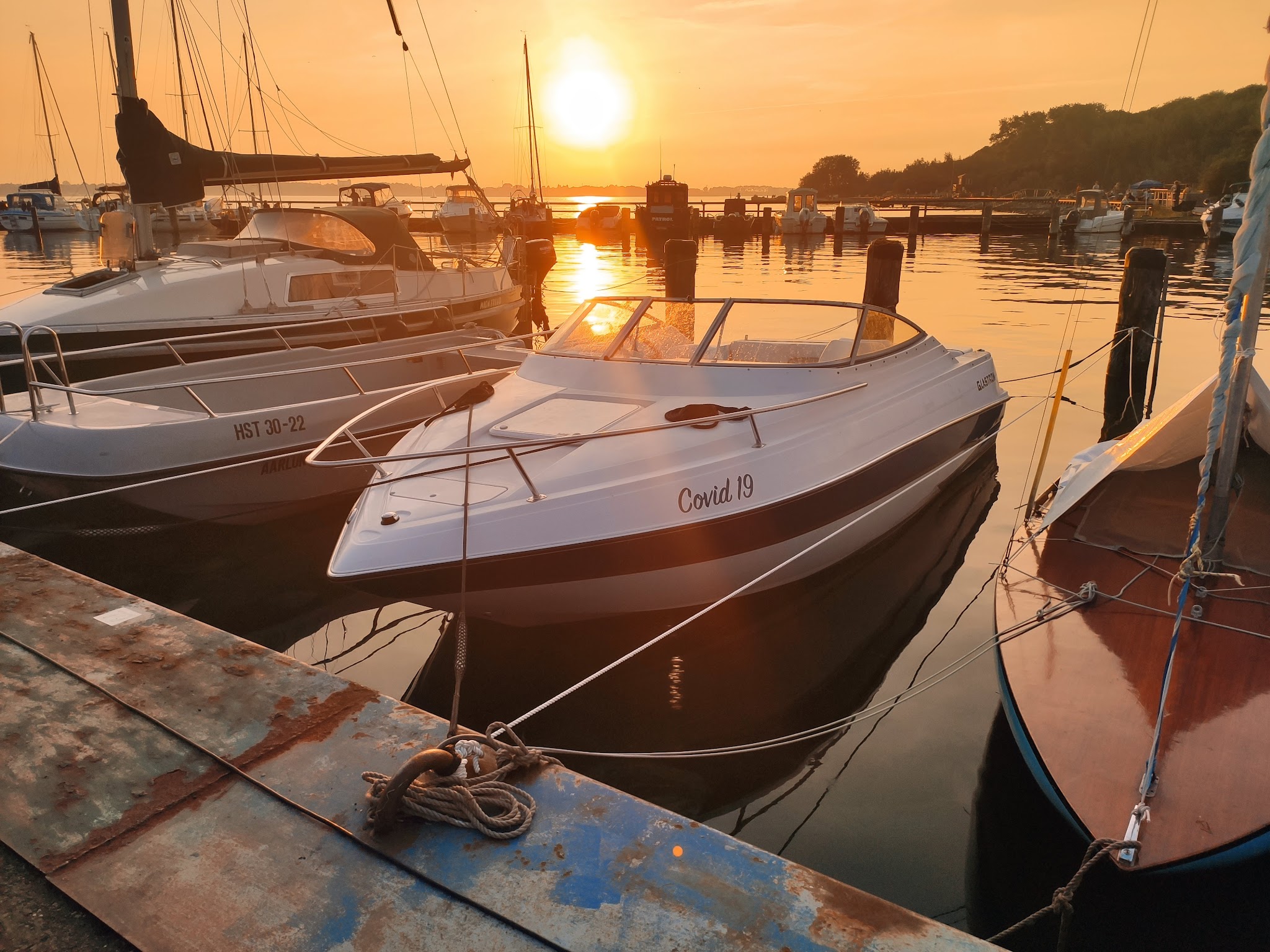 Kleiner Motorboot „Covid 19“ vertäut zwischen Segelbooten, ruhiger Hafen im goldenen Sonnenuntergang