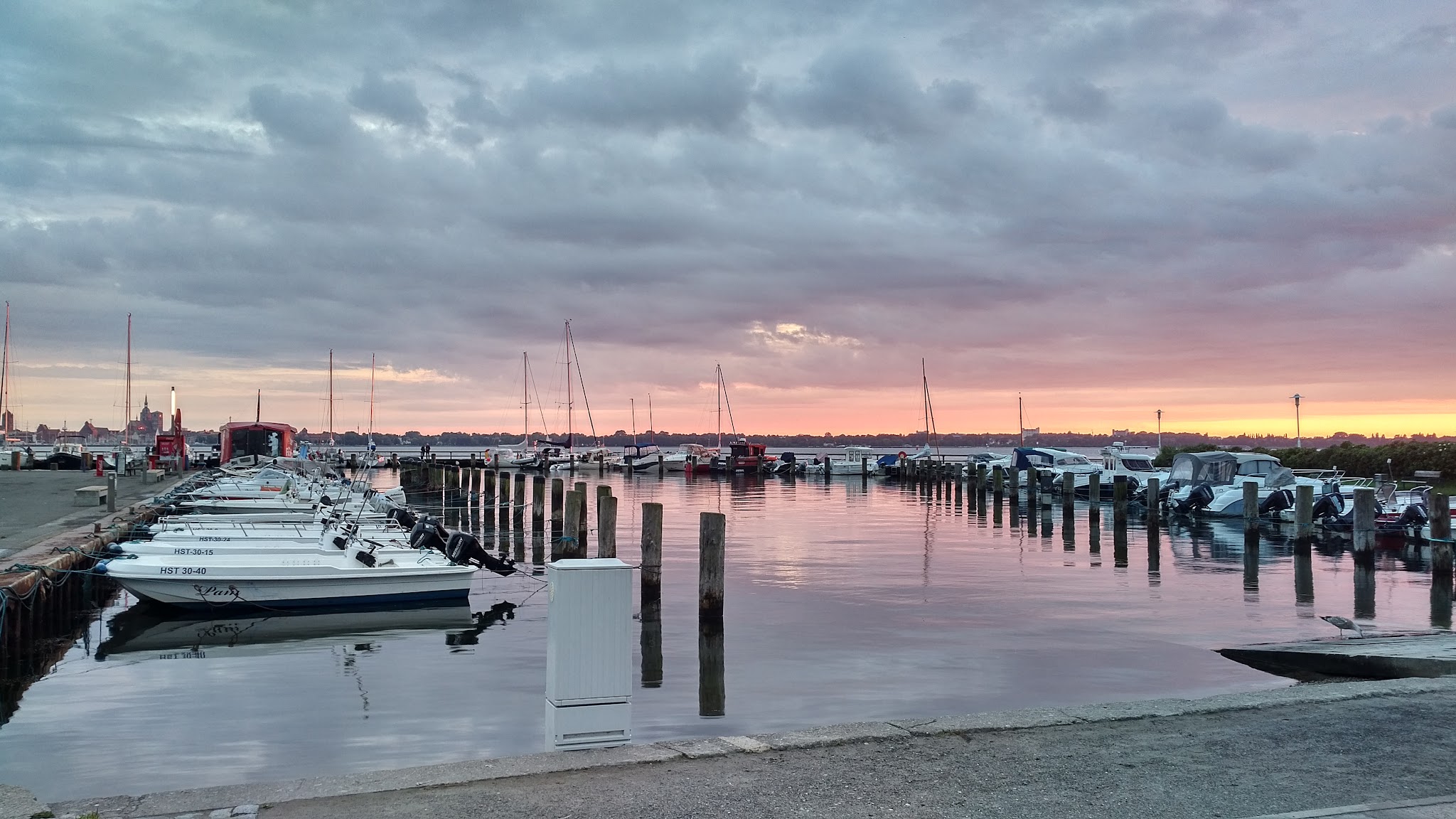 Ruhiger Hafen mit festgemachten Booten; pink-goldener Sonnenuntergang spiegelt sich in stillem Wasser unter Wolken.