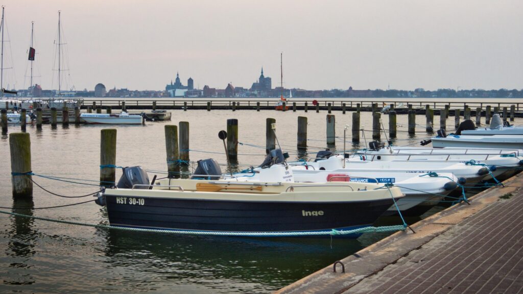 Motorboote am Steg, vorn Boot 'Inge'; ruhiger Hafen mit Holzpfählen, ferne Stadt-Silhouette im Dämmerlicht