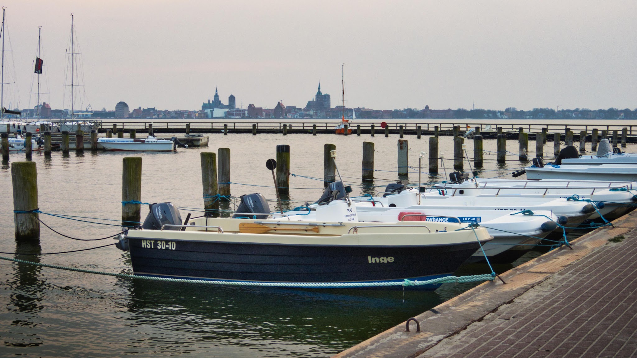 Motorboote am Steg, vorn Boot 'Inge'; ruhiger Hafen mit Holzpfählen, ferne Stadt-Silhouette im Dämmerlicht