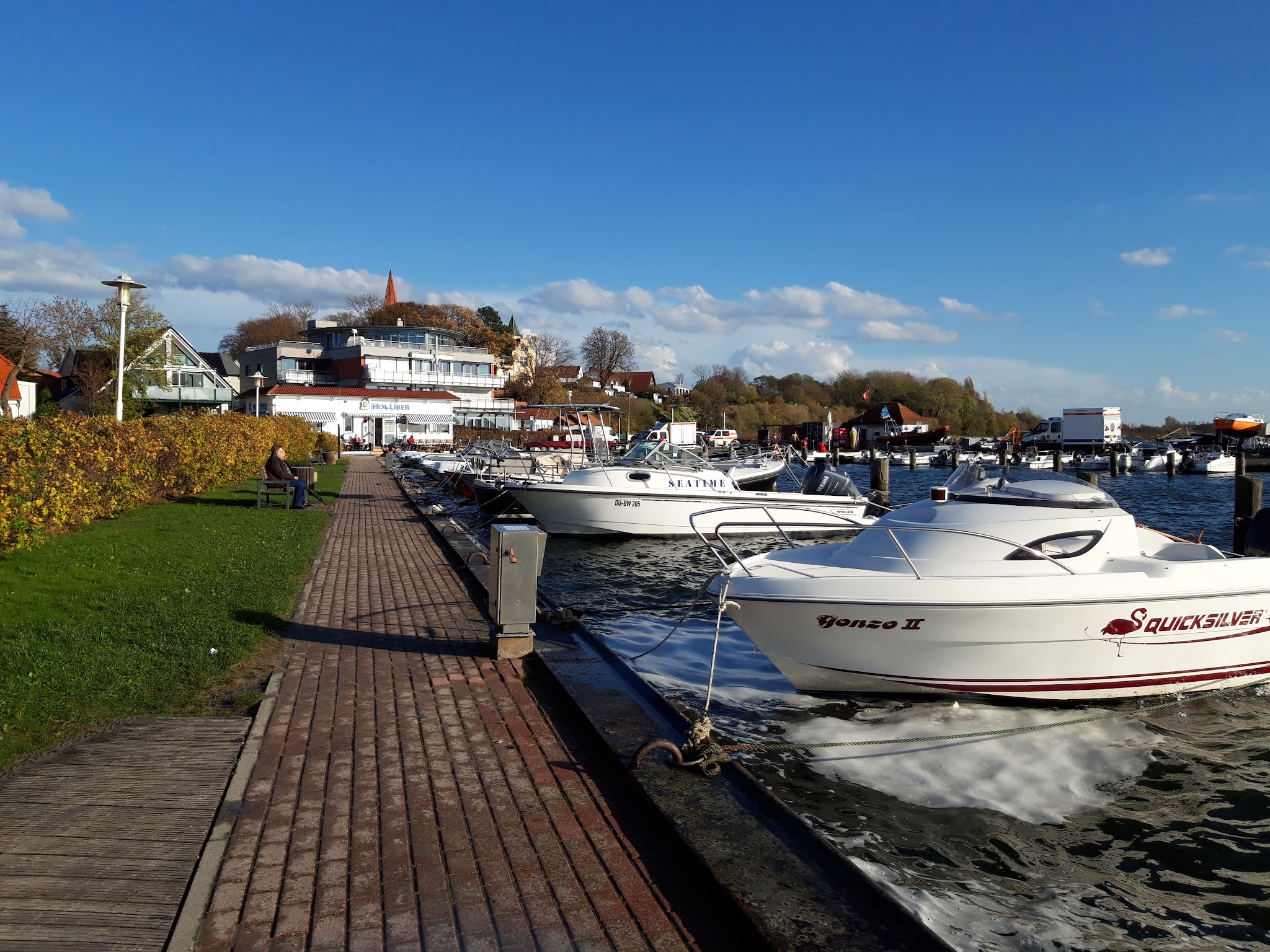 Sonniger Yachthafen mit weißen Motorbooten am Steg, Person auf Bank, modernes Ufergebäude, klarer blauer Himmel