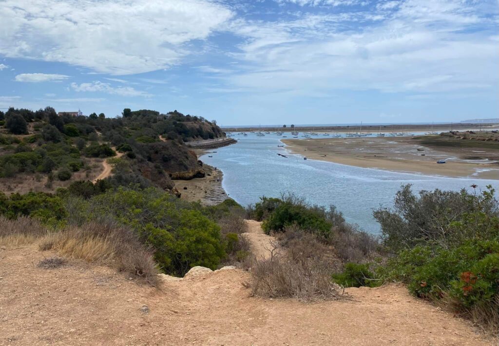 Malerische Flussmündung bei Ebbe: sandige Bänke, viele vor Anker liegende Boote, bewachsener Küstenpfad.