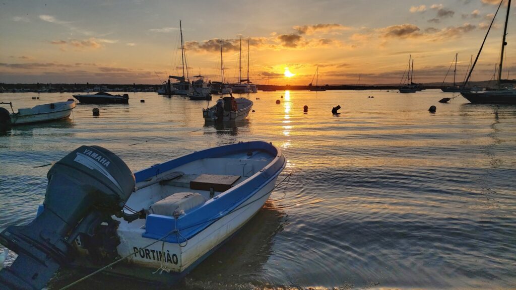 Motorboot im Vordergrund, dahinter ankernde Segelboote auf ruhiger Bucht, goldener Sonnenuntergang spiegelt im Wasser.