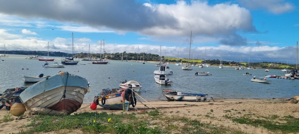 Kleine Fischerboote liegen am sandigen Ufer; weitere Segelboote ankern auf ruhiger Bucht vor grünen Hügeln.