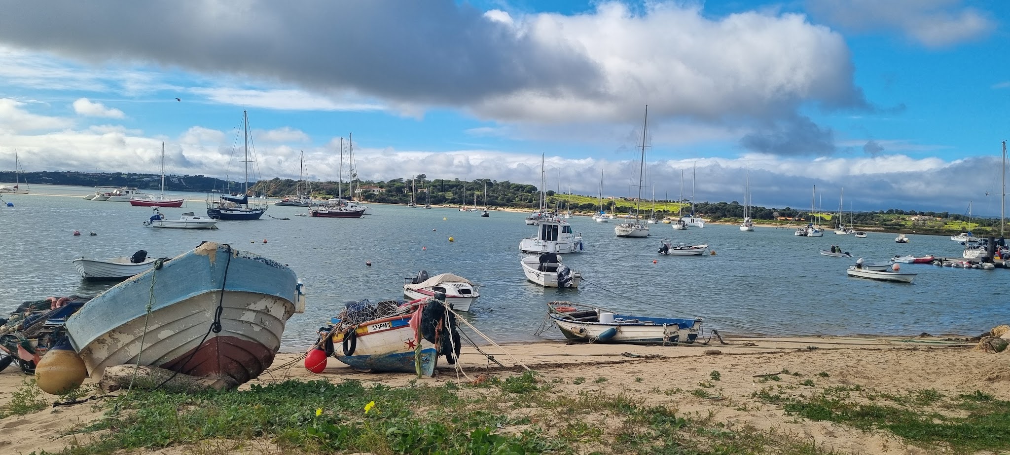 Kleine Fischerboote liegen am sandigen Ufer; weitere Segelboote ankern auf ruhiger Bucht vor grünen Hügeln.