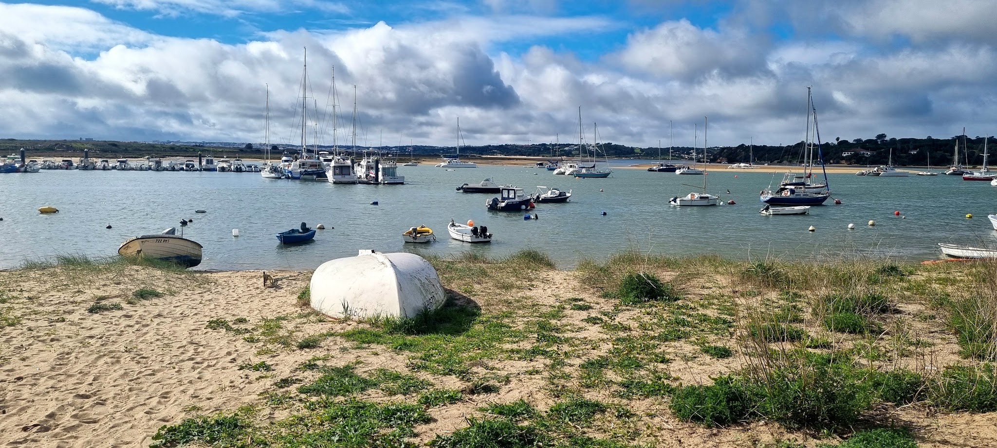 Sandige Uferbucht mit umgedrehtem Ruderboot, dahinter zahlreiche Segel- und Motorboote auf ruhigem Wasser.