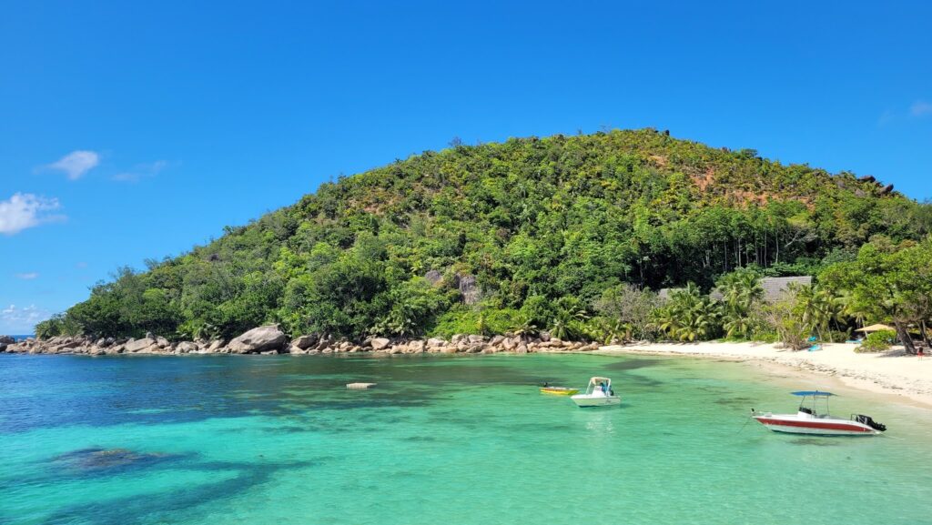 Türkisblaue Bucht mit weißem Sandstrand; dicht bewaldeter Hügel im Hintergrund, zwei kleine Boote auf dem Wasser