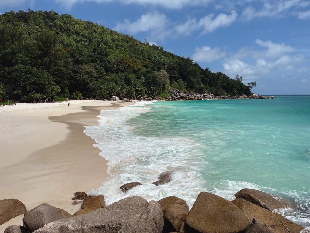 Tropischer Strand mit weißem Sand, türkisfarbenem Meer, dichtem grünen Hügel, Granitfelsen vorn und wenigen Badenden