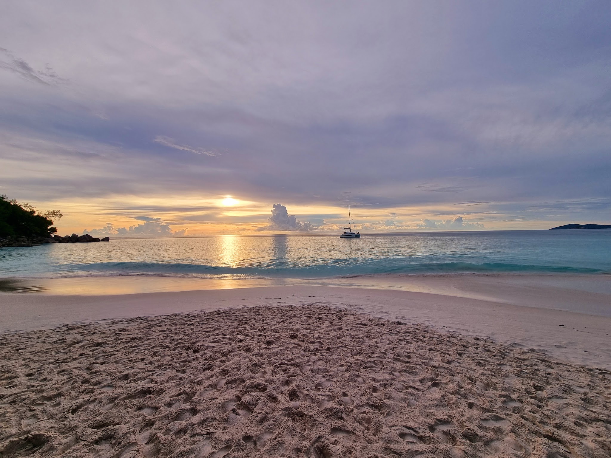 Sonnenuntergang über ruhigem Meer, einsames Segelboot vor türkisblauem Wasser, sandiger Strand im Vordergrund.