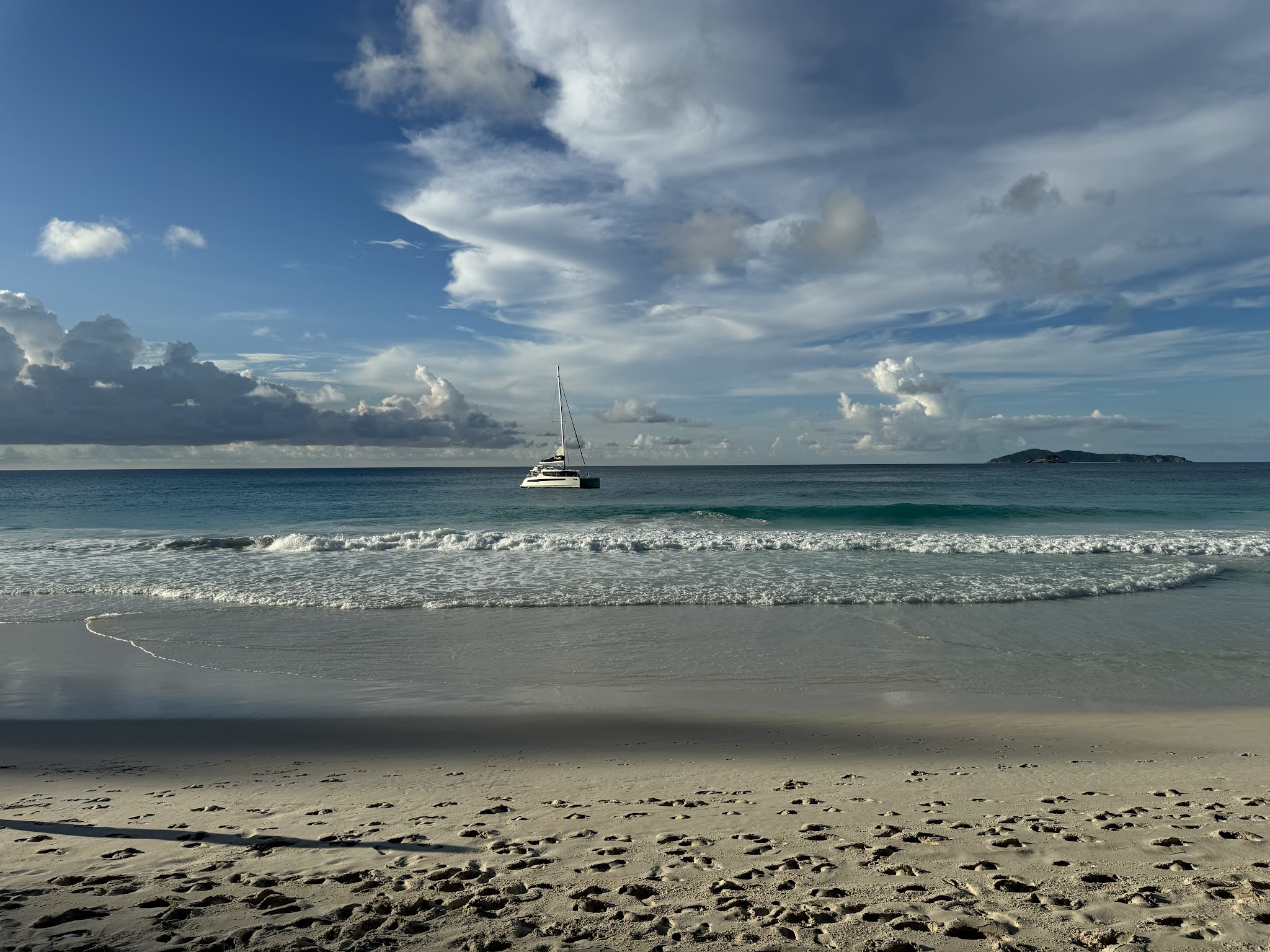 Katamaran vor tropischem Sandstrand; sanfte Wellen, Spuren im Sand und wolkenreicher Himmel mit Insel in der Ferne