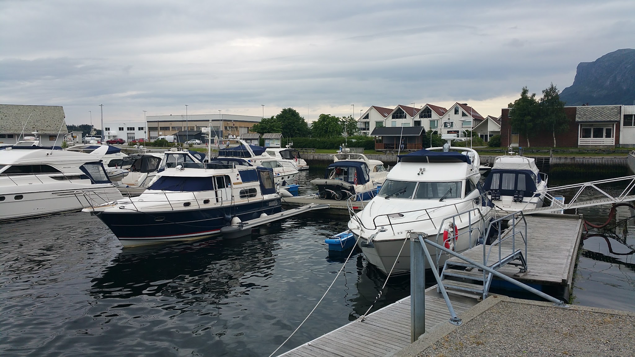 Mehrere Motorboote und Yachten liegen in einem kleinen Hafen vor Wohnhäusern unter bewölktem Himmel.