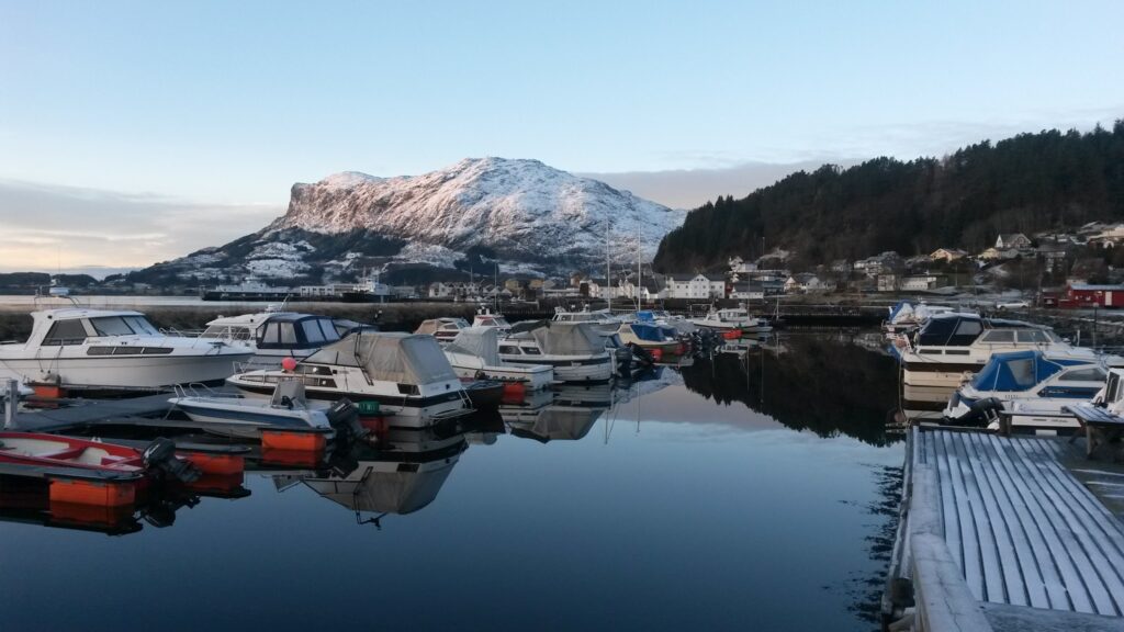 Verschneiter Fjordhafen: ruhiges Wasser spiegelt Boote, Häuser und schneebedeckten Berg im Morgenlicht.
