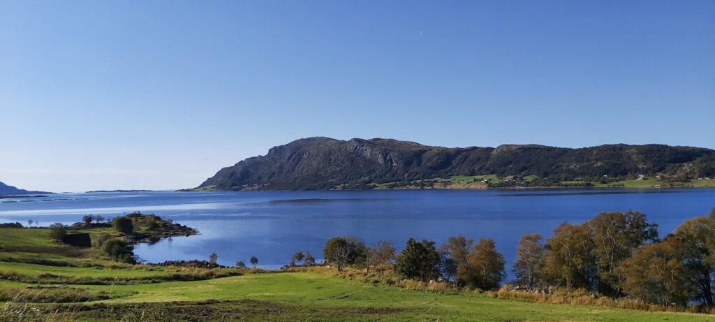 Ruhiger See mit tiefblauem Wasser, grüne Wiesen und Bäume im Vordergrund, dunkler Bergrücken unter wolkenlosem Himmel