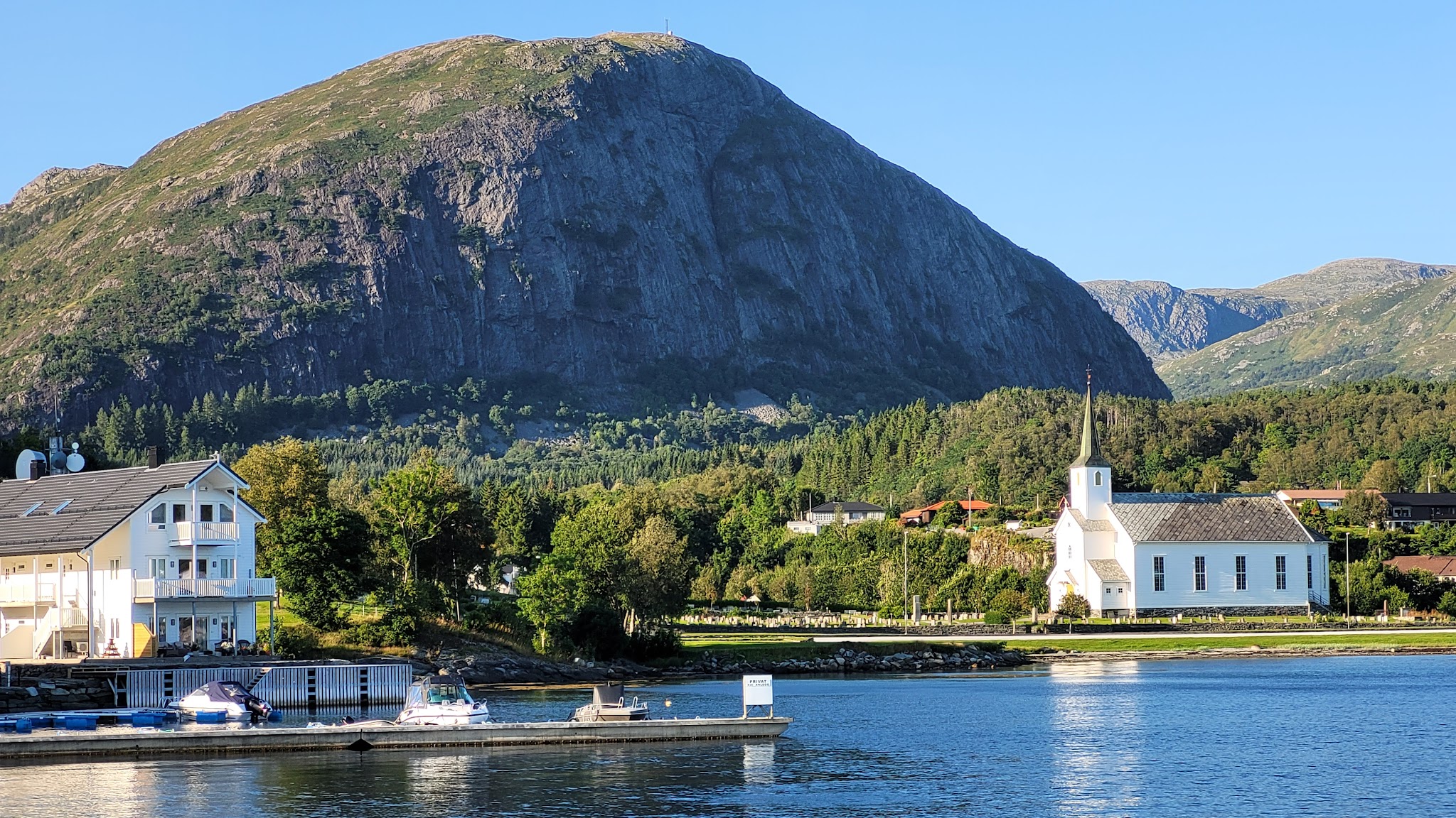 Weiße Kirche und Bootssteg vor Häusern und Wald, dahinter mächtiger felsiger Berg unter wolkenlosem Himmel