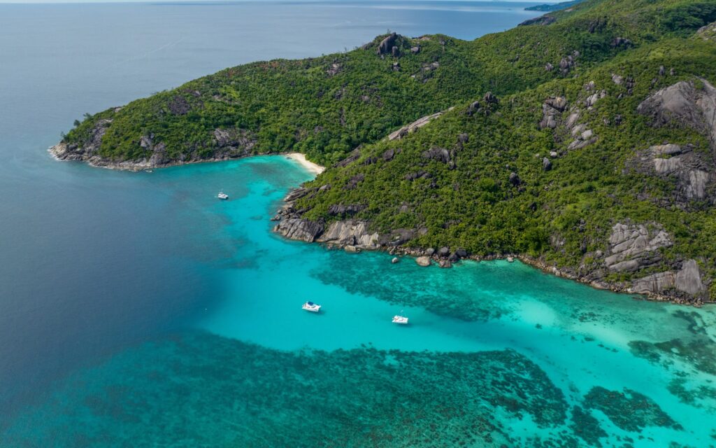 Türkisfarbene Bucht mit Korallenriff, umgeben von dicht bewaldeten Hügeln; mehrere weiße Yachten ankern im klaren Wasser.