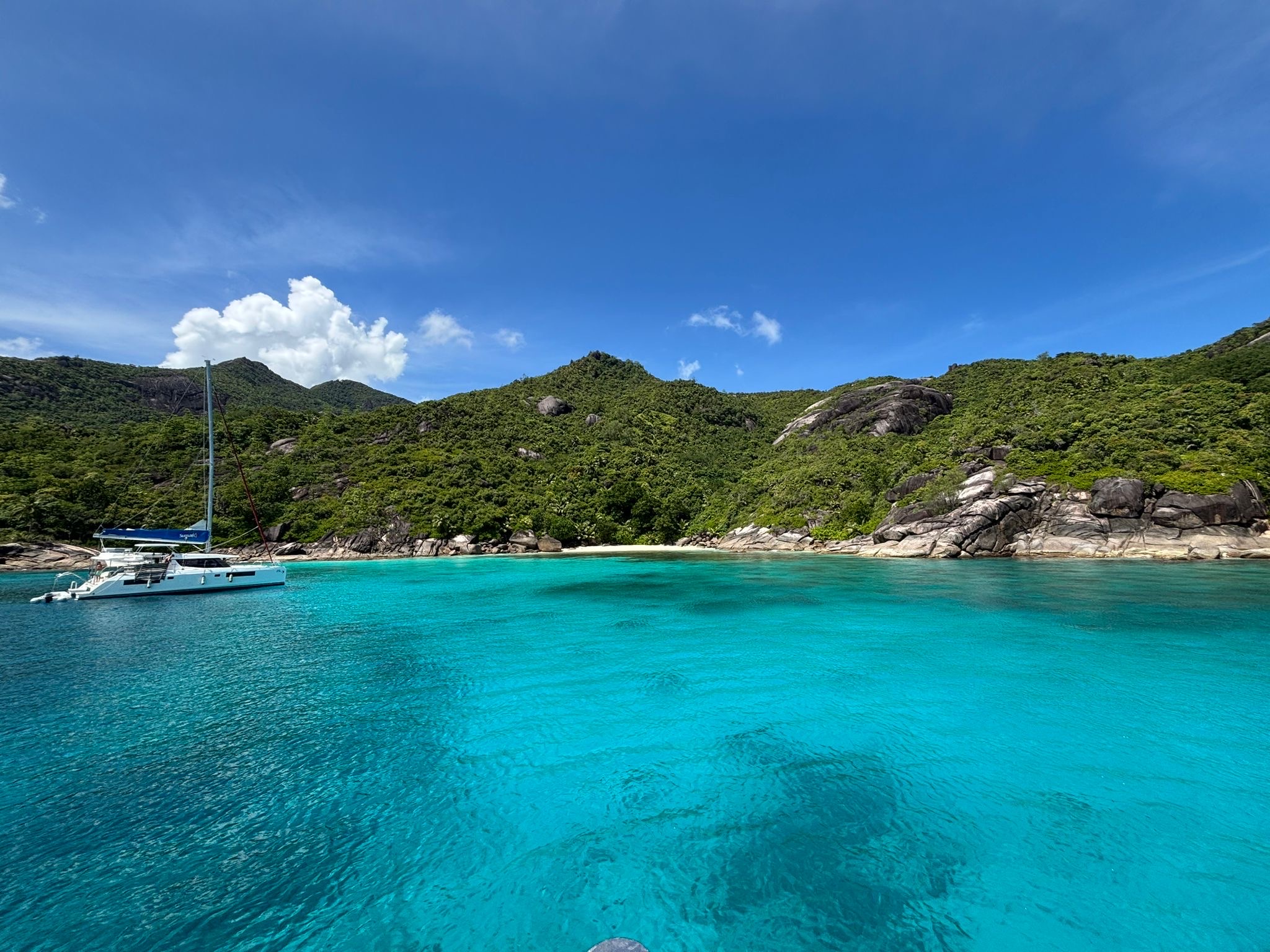 Katamaran ankert in türkisblauer Bucht vor üppig grünen Hügeln und Felsen unter klarem Himmel.