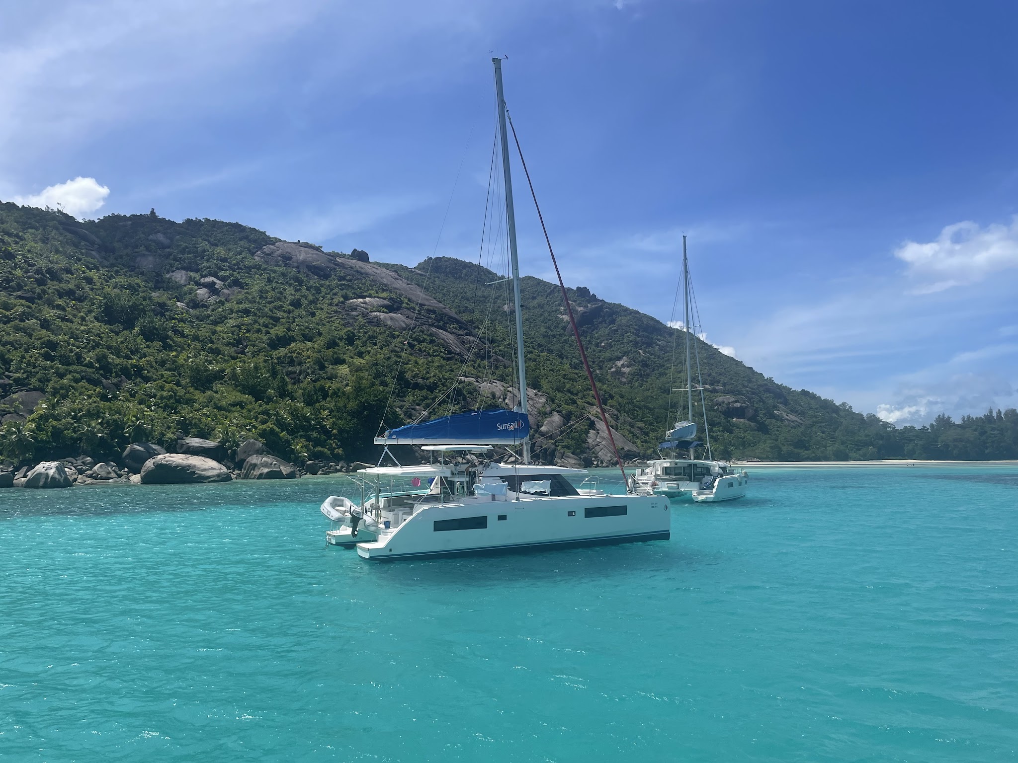 Zwei weiße Katamarane ankern im türkisblauen Meer vor grün bewaldeten Felsen unter klarem Himmel.