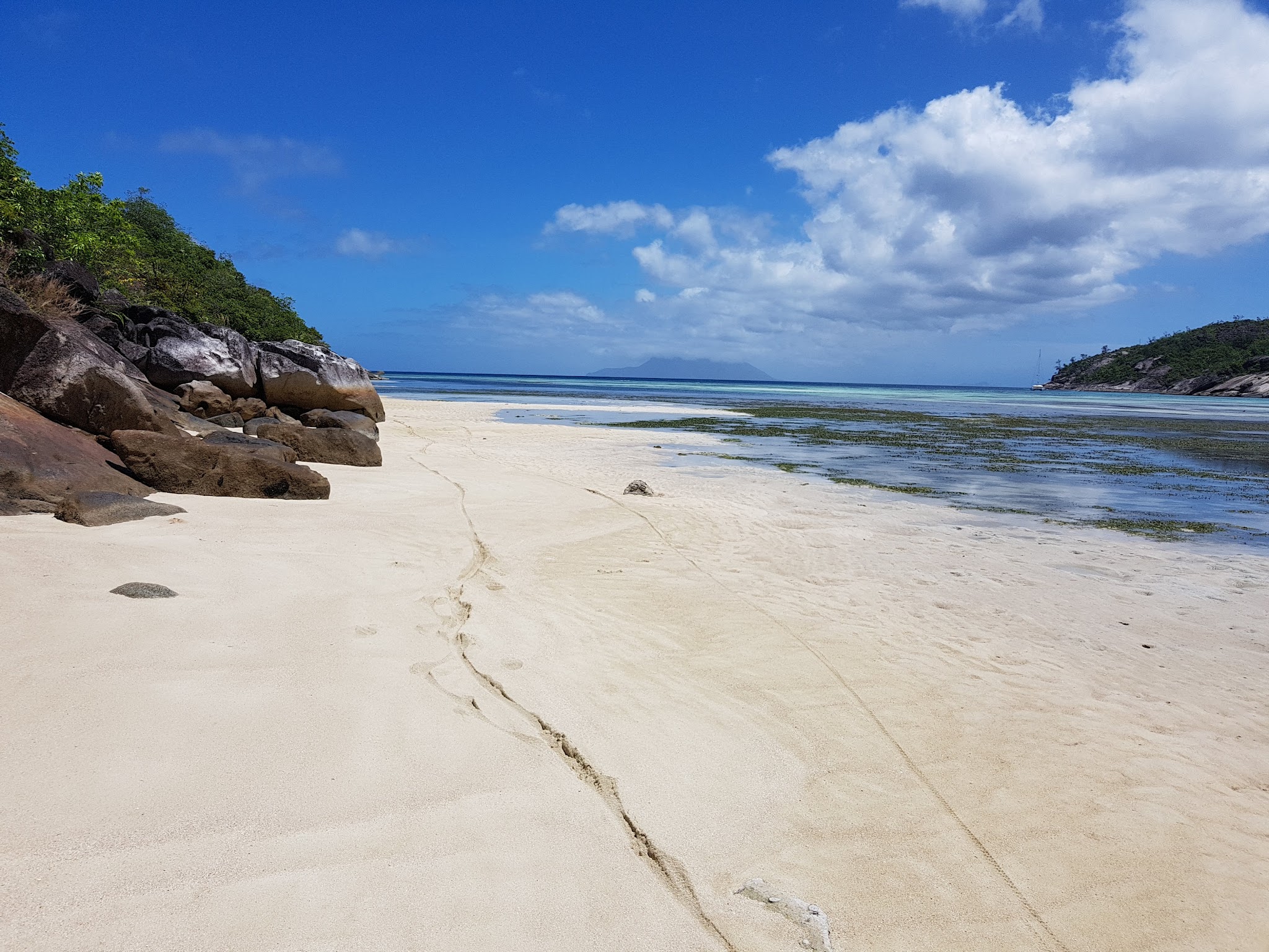 Breiter, heller Sandstrand mit Felsen links, seichtem türkisblauem Meer, Inseln am Horizont unter klarem Himmel
