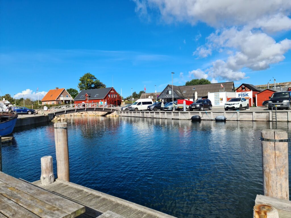 Kleiner Hafen mit klar blauem Wasser, Holzsteg im Vordergrund, bunte Häuser und Autos am Kai unter strahlend blauem Himmel