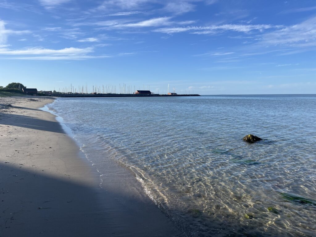Ruhiger Sandstrand, flaches, klares Wasser; im Hintergrund Hafen mit vielen Segelmasten unter blauem Himmel