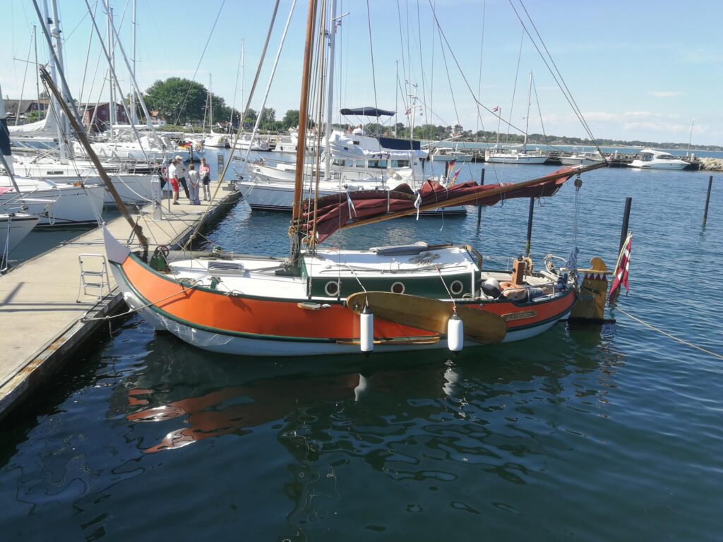 Orange-weißes Segelboot mit dänischer Flagge liegt im Yachthafen; Personen spazieren über den Steg.