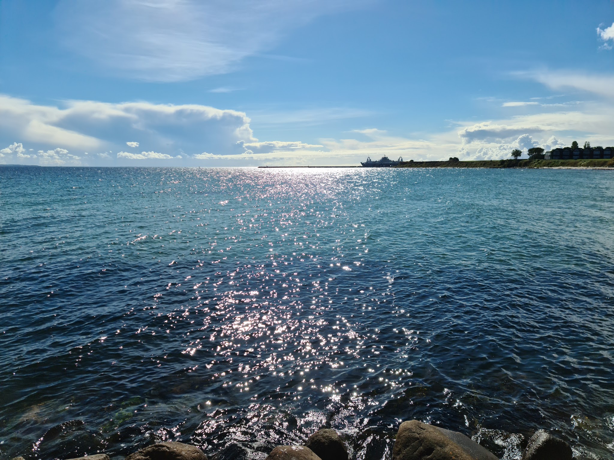 Sonne glitzert auf blauem Meer; im Vordergrund Felsen, am Horizont Küstenlinie mit Silhouette eines Schiffs.