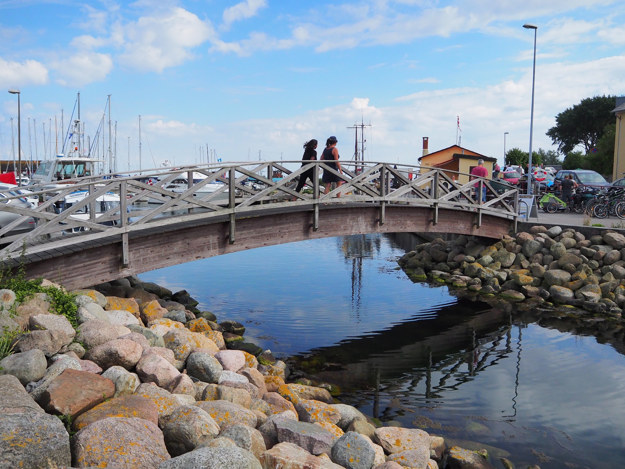 Kleine Holzbrücke im Hafen, zwei Personen gehen darüber; Wasser spiegelt Masten, Felsufer im Vordergrund.