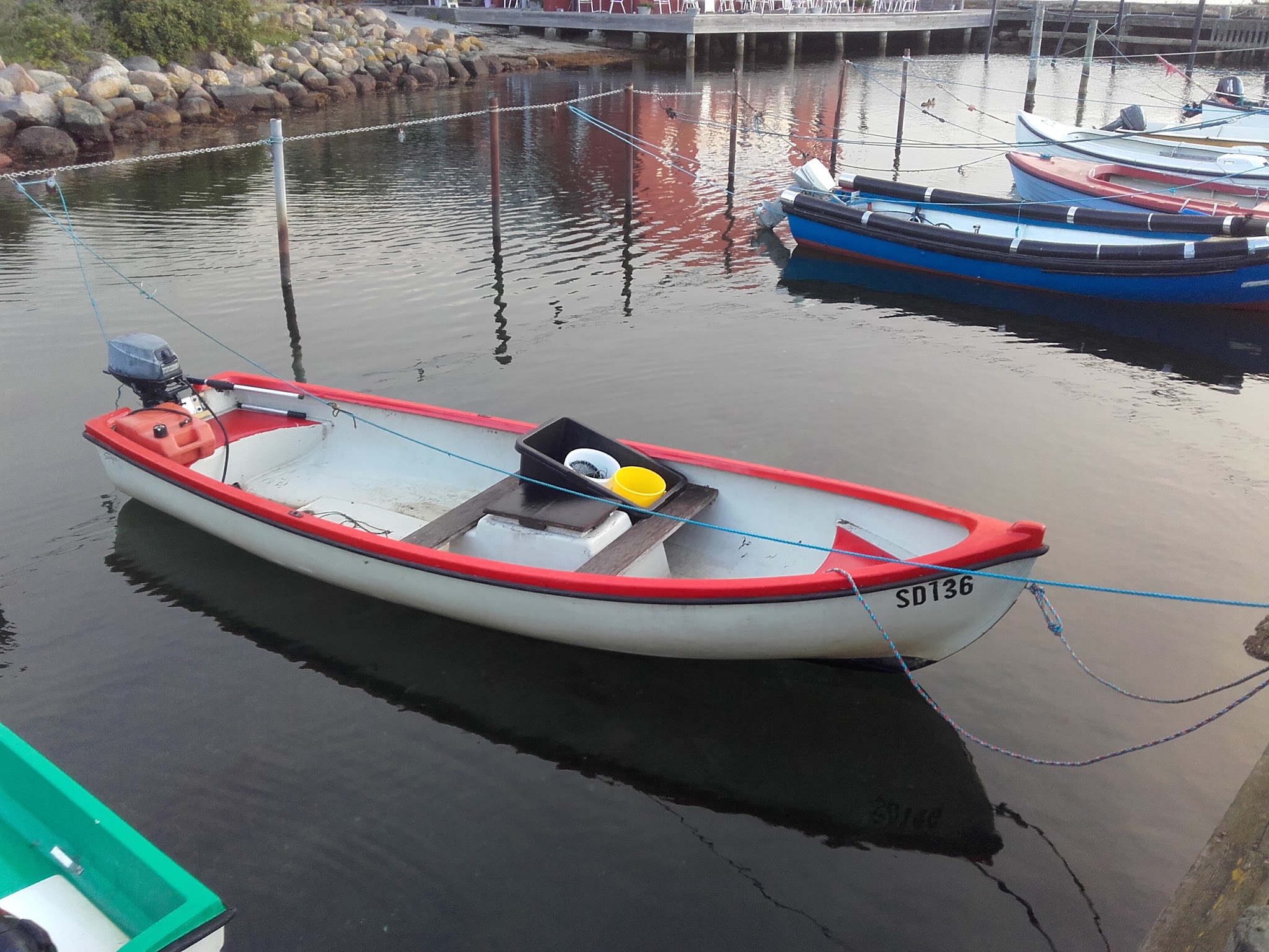 Kleine weiß-rote Motorjolle mit gelbem Eimer, am Anleger vertäut, weitere Boote spiegeln sich im ruhigen Hafenwasser.