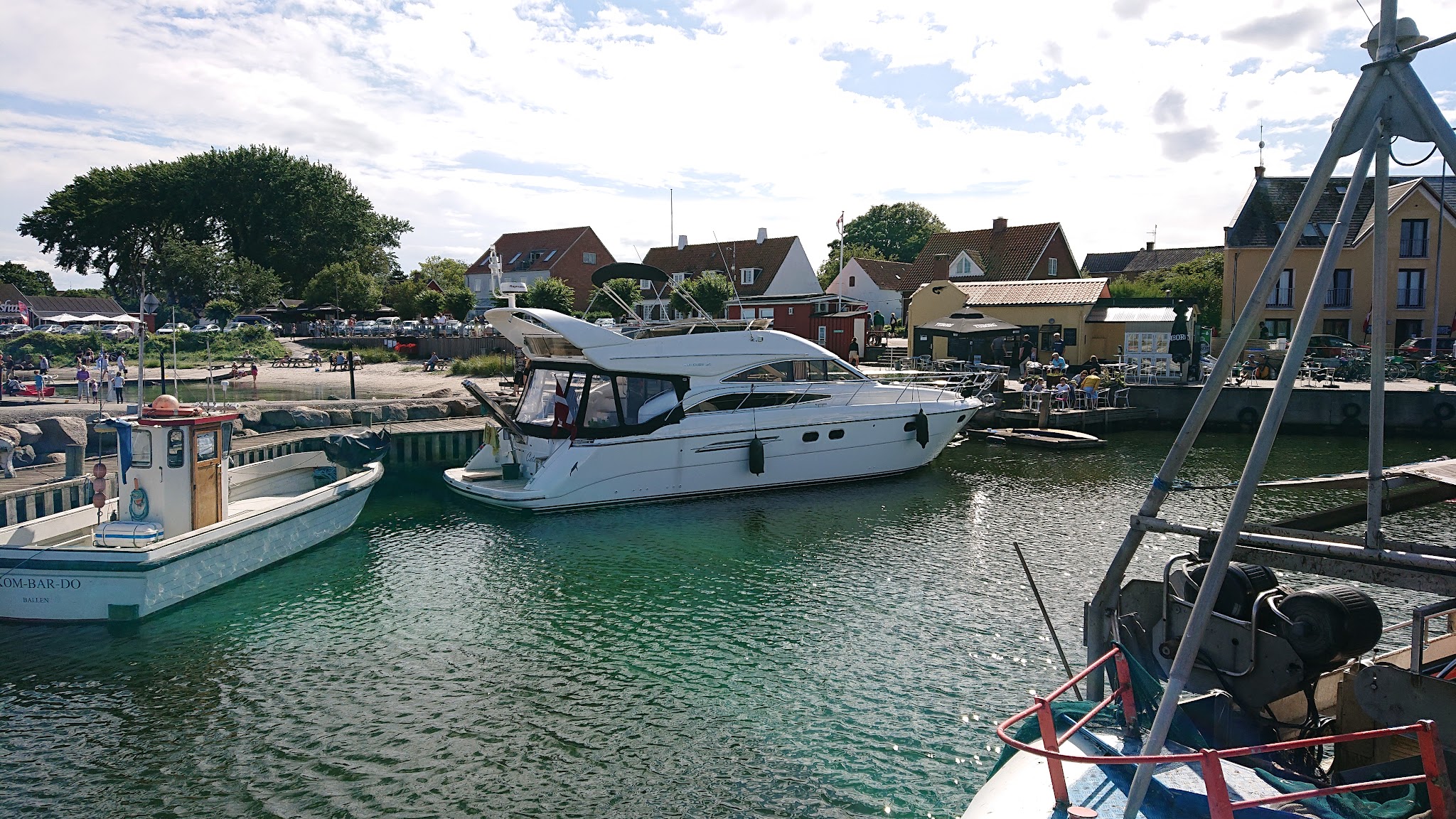 Große weiße Motoryacht liegt im sonnigen Dorfhafen; kleine Boote, Cafés und Häuser säumen das Ufer.