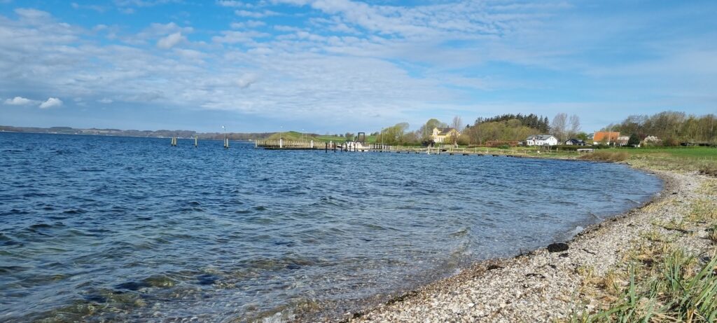 Kieselstrand an ruhiger Bucht, klares blaues Wasser, Steg und vereinzelte Häuser vor grünem Ufer und Wolkenhimmel