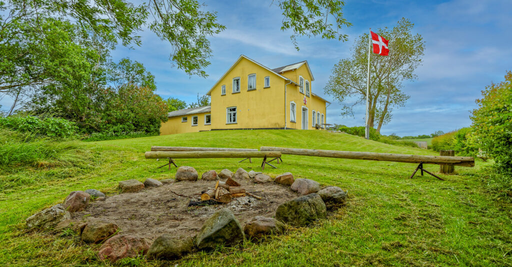 Gelbes Landhaus auf Hügel, davor steinerne Feuerstelle im Rasen; dänische Flagge weht am Mast.