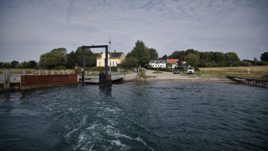Kleine Fähranlegestelle vor Küstendorf; gelbes Haus, ein Auto und Bäume hinter ruhigem Wasser.
