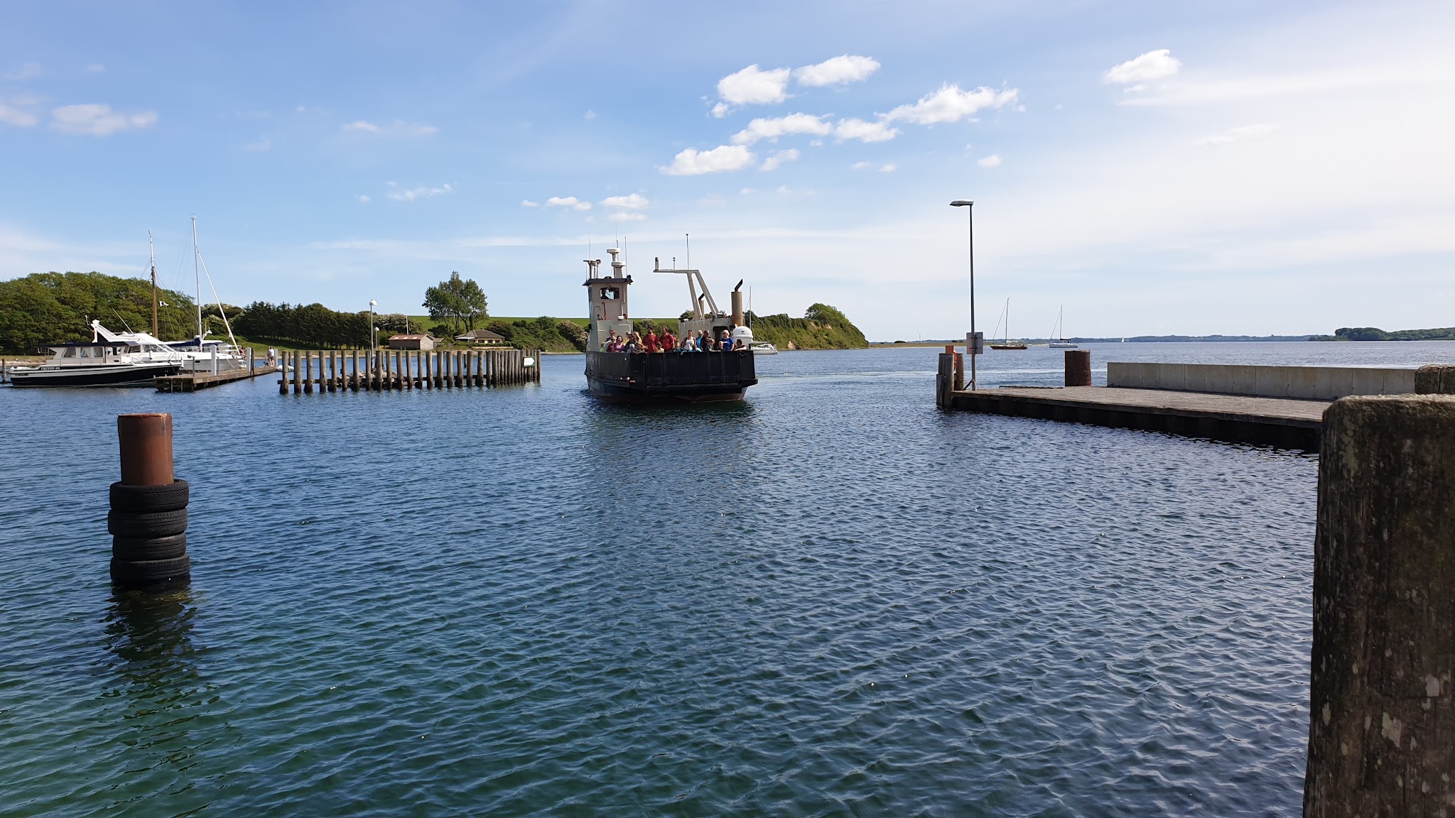 Kleine Personenfähre mit Passagieren fährt an Pier und Segelbooten vorbei in sonniger Küstenbucht.