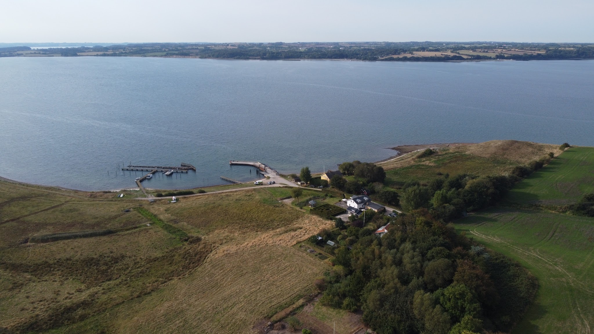 Luftaufnahme einer ruhigen Küstenlandschaft mit Stegen, Wiesen, vereinzelten Häusern und weitem Wasser